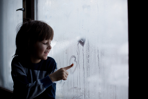 Little boy, leaving finger prints and drawing hearts on a window at home