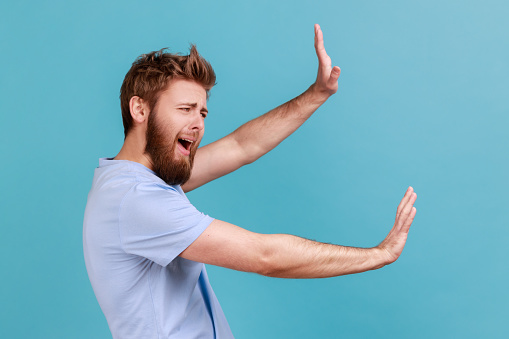 Side view of shocked scared unhappy young bearded man screaming in horror and fear, keeping hands raised to defend himself, panic attack. Indoor studio shot isolated on blue background.