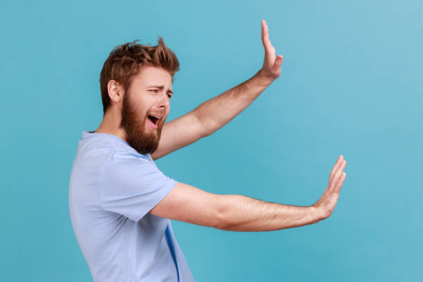 Man in T-shirt screaming in horror and fear, keeping hands raised to defend himself, panic attack. stock photo