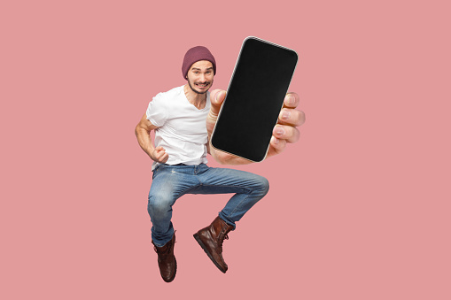 Happy man flying and jumping in air and showing big mobile empty screen for copy space and advertising area. indoor studio shot isolated on pink background