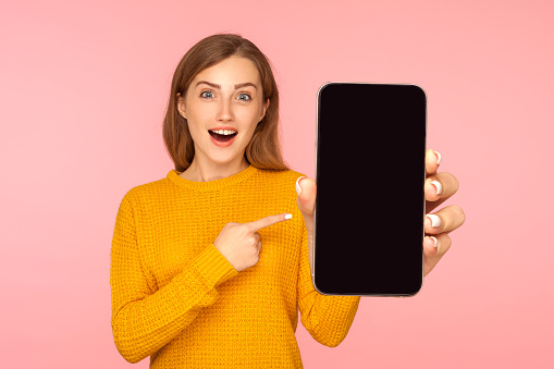 Portrait of amazed good looking girl in sweater pointing at cellphone and looking with surprise at camera, shocked about device or mobile application. indoor studio shot pink background