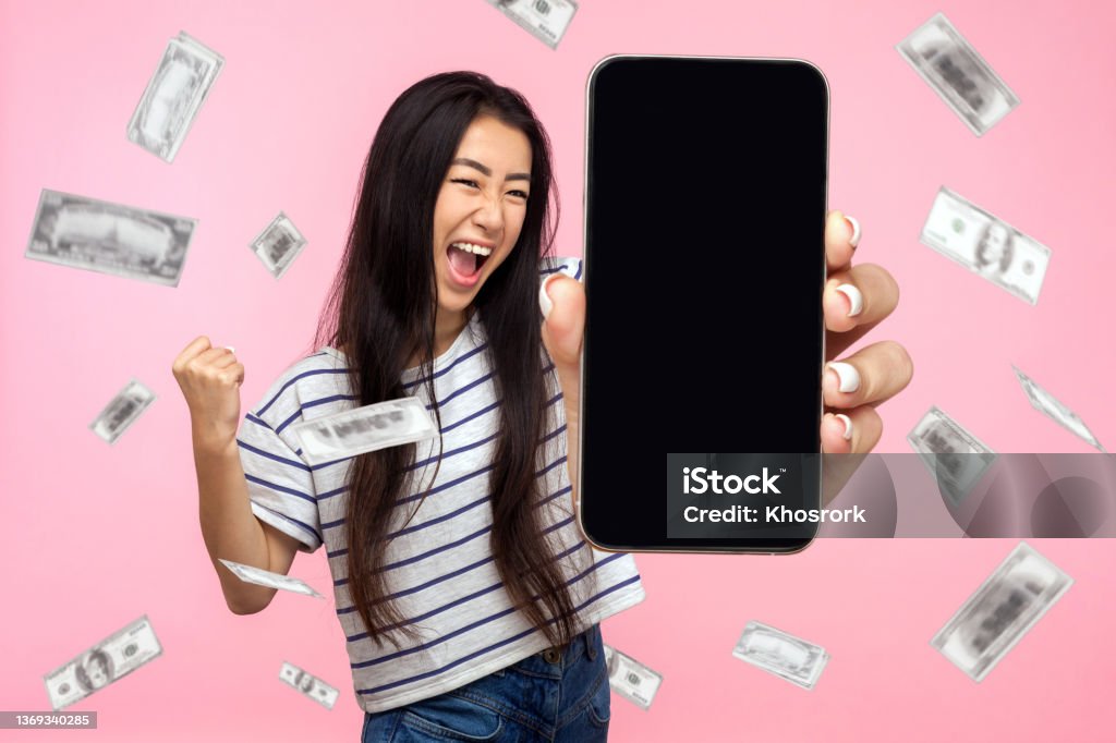 Online investment, winner rich woman and money earning. Online investment, winner overjoyed rich woman and money earning. Portrait of amazed young woman showing mobile empty display and celebrating her victory. indoor isolated on pink background. Lottery Stock Photo