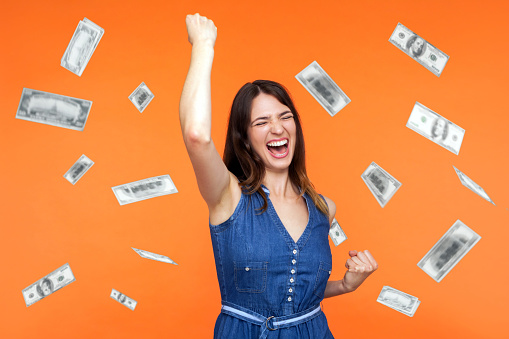 Money rain, winner and rich. Hurray. satisfied happy young woman rising hands up with toothy smile on face, pleased, amazed with money falling. Indoor studio shot isolated on orange background