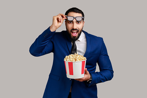 Bearded man being impressed of movie film, hold popcorn, watching amazing movie, keeping mouth open, raising 3d glasses, wearing official style suit. Indoor studio shot isolated on gray background.