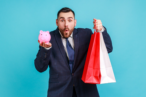 Businessman in dark suit holding paper shopping bags and piggy bank, saving money, cashback, looking at camera with amazed expression. Indoor studio shot isolated on blue background.