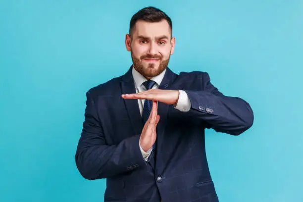 I need more time! Portrait of frustrated brunette man with beard in dark suit showing time out gesture, looking at camera, hurry to meet deadline. Indoor studio shot isolated on blue background.