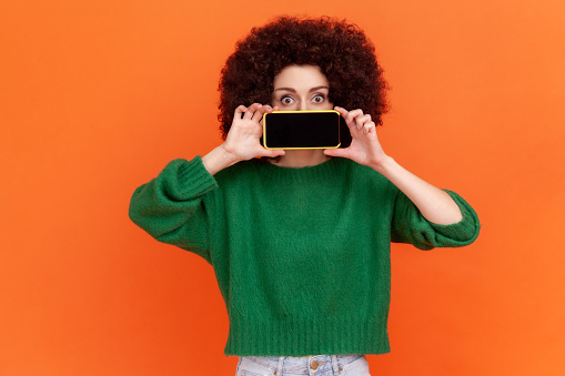 Portrait of woman with Afro hairstyle wearing green casual style sweater covering mouth with smartphone with black blank scree, copy space. Indoor studio shot isolated on orange background.