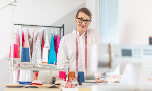 young and confident female tailor stands in the workshop with spools of thread, sewing machine and clothes hangers around her. - seam needle textile industry thread imagens e fotografias de stock