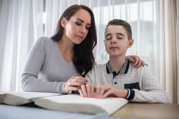 Woman helps visually impaired boy in dark glasses reads from a book in braille. boy to read a book in braille.