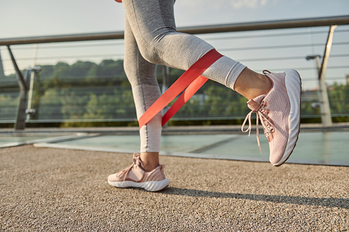Close-up of the legs of an athlete woman doing body weight training with a resistance fitness elastic band on a city bridge.