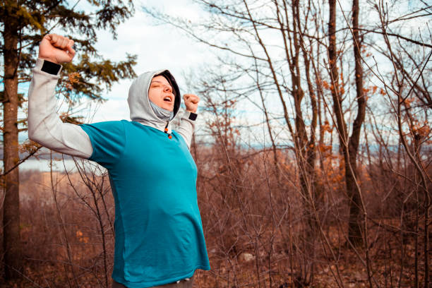Young hooded Man have difficulty breathing while exercise outdoors in cold autumn day in the park.  Stretching his arms and lungs for more air. Copy space