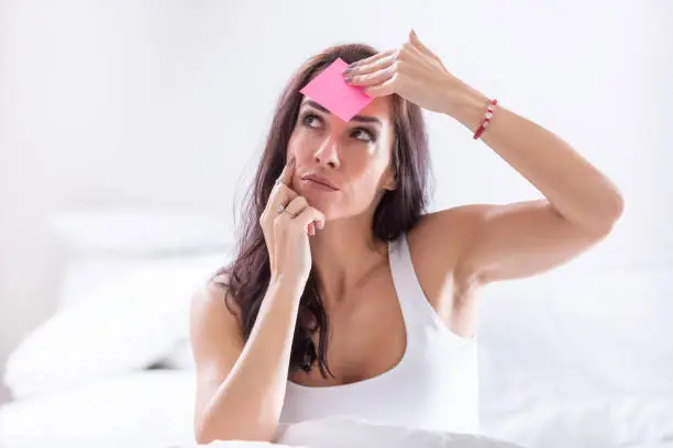 Photo of Woman forgetting something puts a pink post-it on her forehead, thinking.