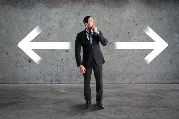 istock Thoughtful young businessman standing on abstract concrete background with two arrows. Different direction and choice concept. 1369355106