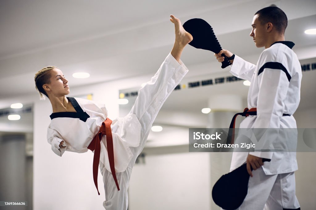 Determined martial artis with disability exercising high leg kick with her coach during taekwondo training. Taekwondo fighter with para-ability practicing high leg kick with her sparing partner at martial arts club. Athlete with Disabilities Stock Photo