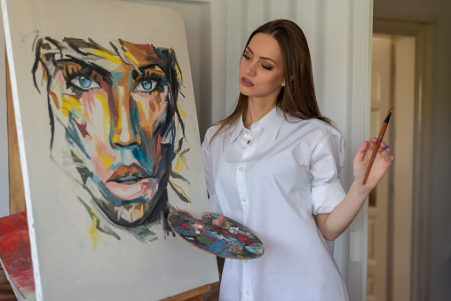 Beautiful female artist painting at home holding artist palette and wearing white shirt