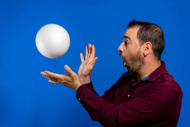 Latin bearded man dressed in a purple shirt with a cork ball isolated on blue studio background, he is throwing the ball imitating Son Goku from dragon ball one of his youth heroes