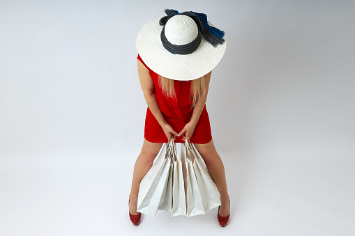 Top view of a blonde in a red dress and a straw hat holding shopping bags on a white background. Girl in red shoes on sale, gets discounts, make purchases