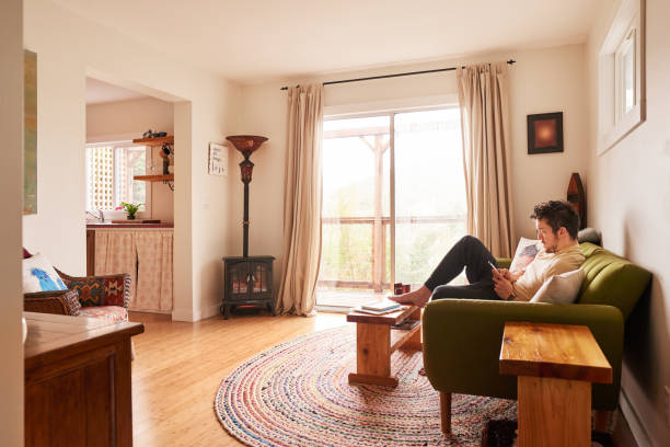 Young man in pajamas sitting on a sofa in a living room in the morning and text messaging on his phone