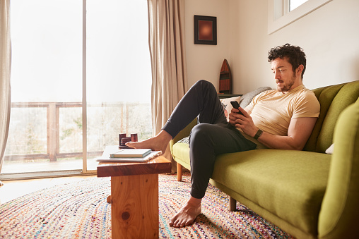 Young man in pajamas relaxing on a living room sofa in the morning and text messaging on his phone