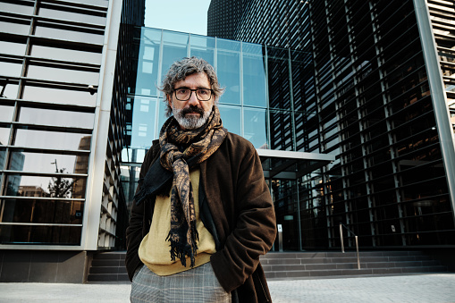 Middle-aged man looking at the camera while posing in the financial district with an office building on background. Business and urban lifestyle concept.