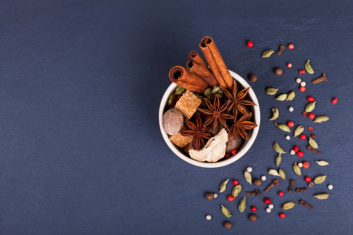 Spices cinnamon sticks, cardamom pods, star anise, sugar, nutmeg, coriander and pepper seeds in brown ceramic cup on a black slate stone plate. Spices for Masala tea.