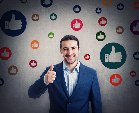 Contented businessman smiling broadly shows thumb up gesture, surrounded by multicolor like symbols. Business project approval and positive feedback
