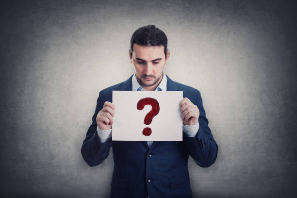 istock Doubtful businessman holding a paper sheet with question symbol, isolated on grey wall background. Depressed person looking down pensive. Interrogation mark as metaphor for business troubles 1369393612