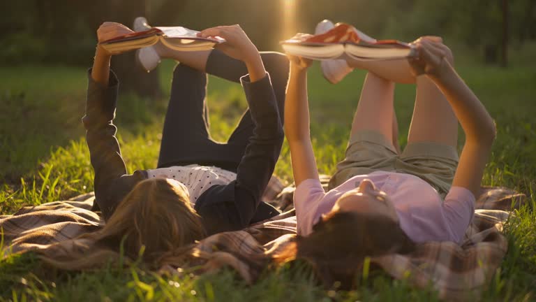 Two relaxed teenage girls lying in sunbeam in summer spring park reading books. Live camera moves left to right as confident concentrated Caucasian friends enjoying leisure at golden sunset.