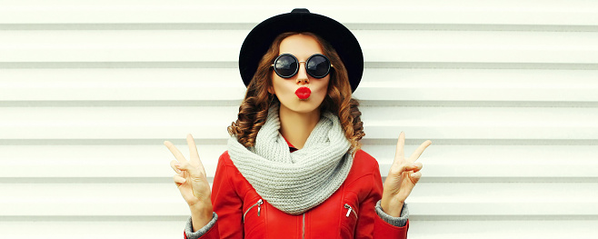 Portrait of stylish woman model blowing her lips sending sweet air kiss wearing scarf and red jacket, black round hat on white background