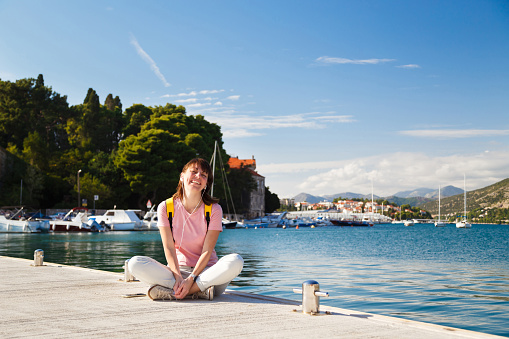 Smiling young woman sitting cross-legged on pier, listening to music with Croatian coastline in background. Bright sunny day in Dubrovnik, Croatia. Travel lifestyle concept