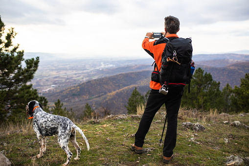 A senior man is taking  picture of the view while hiking with his dog in the woods in autumn