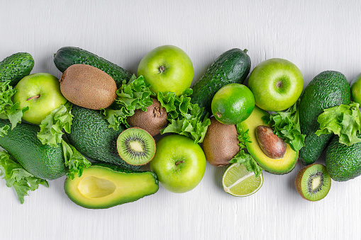 Different green fruits and vegetables whole and sliced such as avocado, apple, lime, kiwi, lettuce and cucumber laying on white wooden background used as ingredients for healthy eating and vegan diet