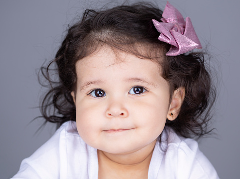 Adorable one year old baby girls headshot, she is looking directly to the camera smiling. She has a white top, the background is gray and her hair is long.