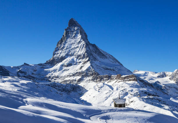 Riffelberg, Switzerland - January 06. 2022: the small Riffelberg chapel Brother Klaus stands on the snow-covered hill with the giant Alps summit Matterhorn at the background.