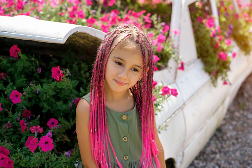 A cute girl with pink afro-pigtails and a green dress is standing by a blooming flower bed with petunias. Colored long pigtails of a child made of artificial hair