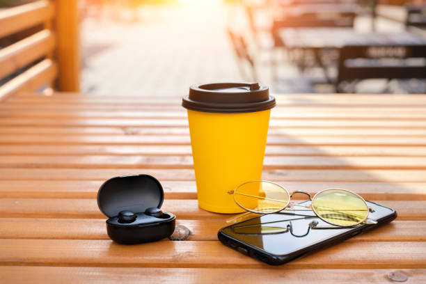 A smartphone, wireless mini earbuds in a case, a disposable cup of coffee and yellow sunglasses are lying on the table of the Gordian cafe on the terrace outside.