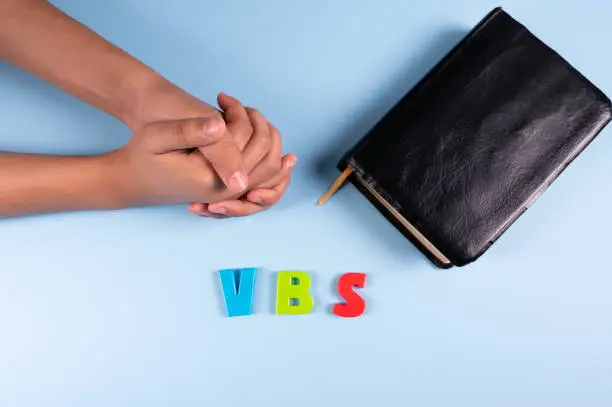 Female hands is praying next to black bible and Abbreviation "VBS" (Vacation Bible School). Copy space text. Selective focus.