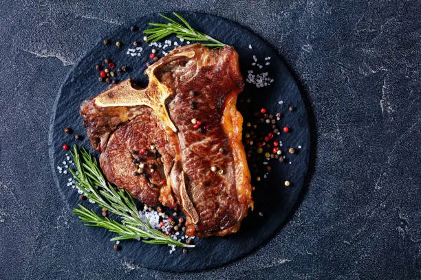 fried porterhouse steak, t-bone steak with thyme, rosemary and peppercorn on black plate on concrete table horizontal view from above, flat lay, free space