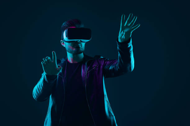 Serious young man in trendy leather jacket and modern VR glasses experiencing virtual reality in dark room with neon illumination