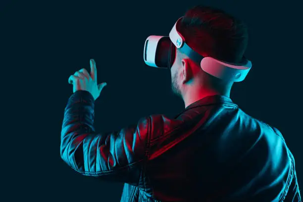 Back view of male player touching invisible screen while experiencing cyberspace, in VR glasses on dark background in studio with pink and blue neon lights