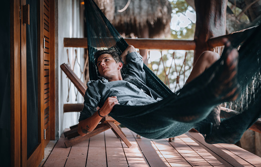 A young urban man relaxing in a hammock hanging on the balcony of a beautiful eco wooden house