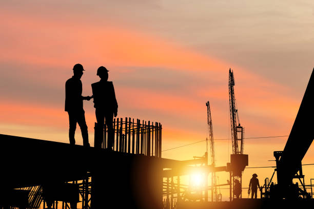 istock Silhouette of Engineer and worker team checking project at building site background, Construction of a mass transit train line in progress with heavy infrastructure site at sunset in evening time 1369474393