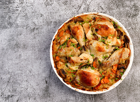Oven Baked Chicken and Rice with onions, carrots and herbs in a white baking dish on a dark grey background. Top view, flat lay