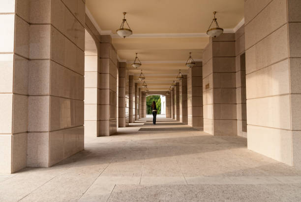 Luanda, Angola: long, elegant colonnade with a series of evenly spaced, arched openings that create a sense of symmetry and perspective. Hanging from the ceiling are large, ornate black lanterns. In the far distance, two silhouetted figures walk side by side, adding a sense of scale and depth to the image - shade along Luanda's 'Avenida Marginal', the corniche.