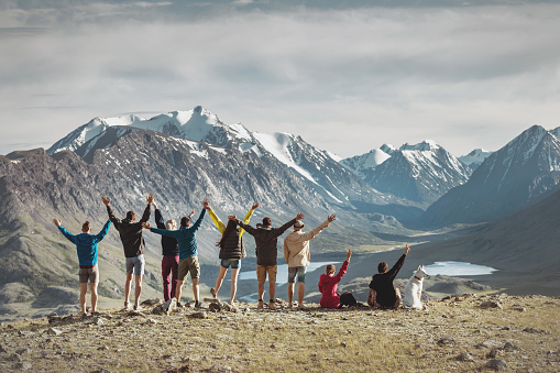 Big group of happy young hikers are standing with raised arms in mountains