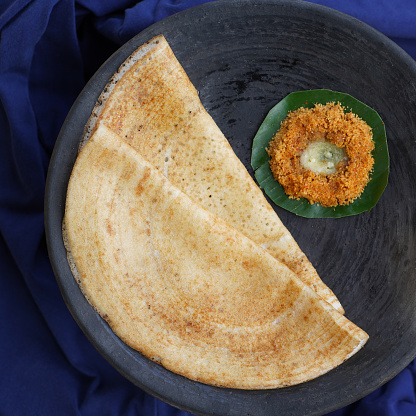 South Indian snack Dosa or dosai or thosai made with rice, black gram or urad dal served on a banana leaf kept on black earthern plate with orange malgapodi or milagai podi or gun powder chutney and ghee or clarified butter.