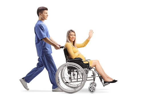 Full length profile shot of a male nurse pushing a pregnant woman waving and sitting in a wheelchair isolated on white background