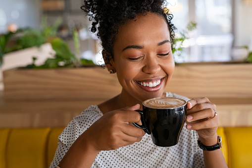 Smiling black young woman smelling freshly brewed coffee with eyes closed in modern cafeteria. Beautiful african american girl in casual clothing smelling and smiling while relaxing in a coffee shop. Close up face of young girl smelling latte coffee in mug at snack bar after university lectures.