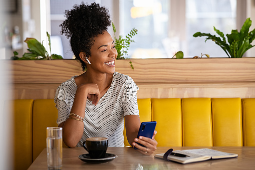 Smiling young woman with earphones holding phone and drinking coffee in modern cafe while looking away. Cheerful black woman using mobile phone and wireless earphones while doing a call in cafe. Positive beautiful girl sitting and relaxing in coffee shop.