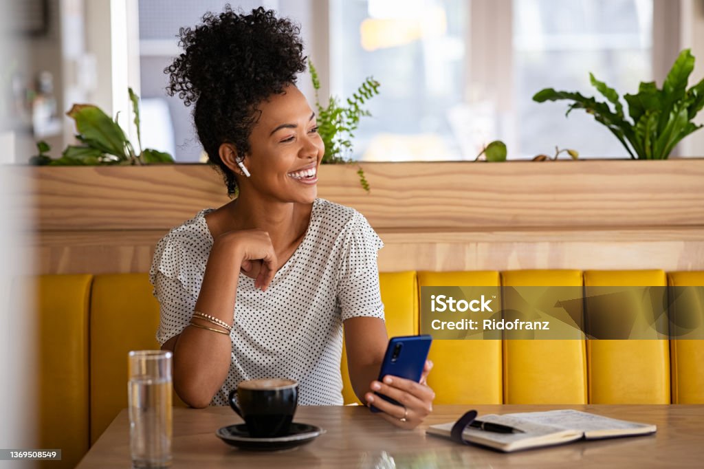 Happy woman in cafe talking on the phone with earphones Smiling young woman with earphones holding phone and drinking coffee in modern cafe while looking away. Cheerful black woman using mobile phone and wireless earphones while doing a call in cafe. Positive beautiful girl sitting and relaxing in coffee shop. Women Stock Photo